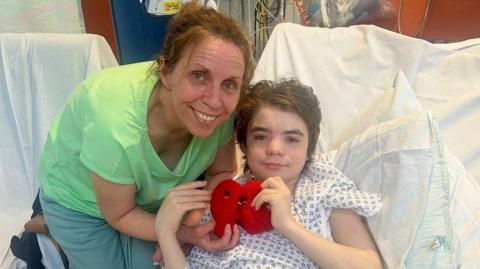A woman wearing a green shirt and pale green trousers, smiles as she stands next to a boy with dark hair who is in a hospital bed wearing a gown. They are both holding cuddly toy kidneys. 
