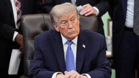 U.S. President Donald Trump sits at his desk in the Oval Office turning to his left. He is wearing a blue suit, white shirt and blue tie. He is not smiling. 