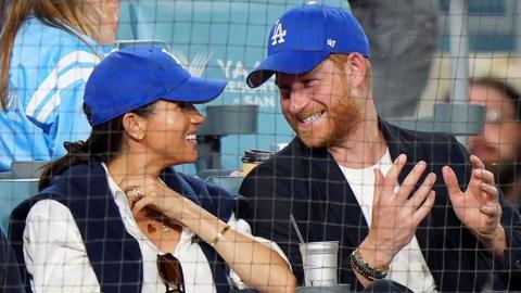 Meghan Markle and Prince Harry wear blue Dodgers baseball caps while seated in the front row during the third inning between the Toronto Blue Jays and the Los Angeles Dodgers during Game 4 of the 2025 MLB World Series at Dodger Stadium.