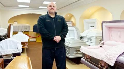 John Redhead standing in a funeral directors with dozens of coffins around him