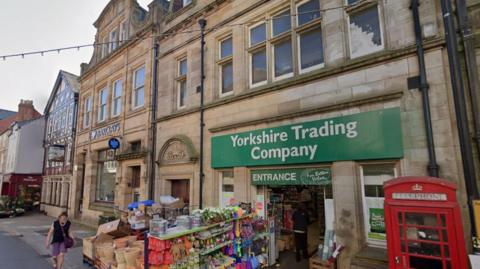 Stone buildings on a shopping street. The closest has a green sign over the door saying Yorkshire Trading Company. Outside it is a red public telephone box and stands stacked with goods including plant pots. Next door is a building with a blue Barclays sign above the window. 