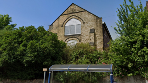 The view of Rock House from Barnsley Road, a disused building with a tall, triangular roof. The front face of the building is made from stone masonry, but the rest of the walls are made from red brick.