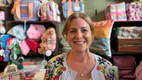 Harriet Rouse smiling towards the camera in her shop. She is wearing a stripy and floral jacket and is wearing a necklace. In the background there are colourful items on shelves. 