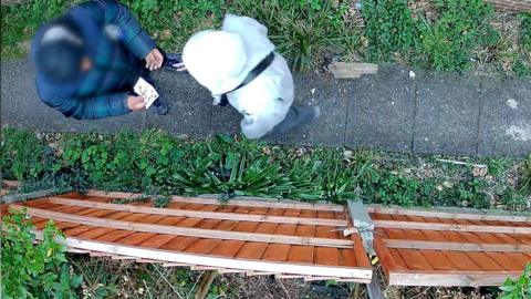 A still of CCTV footage which looks down on two figures - one of whom appears to be holding bank notes. They stand on a path surrounded by shrubbery with a brown fence on one side. The individual on the left is blurred and you cannot see either individual's face.