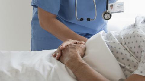 A stock image of a person in a hospital bed with medic stood next to them in blue scrubs.