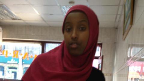 Zahwa Salah Mukhtar in a red headscarf and black top. She stands inside a building with cream tiles on the walls and a window at the back. 