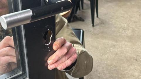 A man is changing the lock in the shop's front door. Through the glass the man can be seen, he has a ginger beard and moustache.