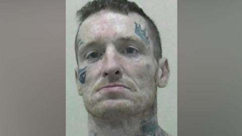 Mugshot of Gary Haswell. He has short dark hair and a faint ginger stubble beard. He has various tattoos, including of a large crown above his left eye, a tribal mark around his right eye and stars and large flowers across his neck.
