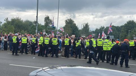 A line of 15 police officers standing in a road as cars pass in the foreground, with their backs to the camera as they face a large group of protesters, some of whom are waving flags. There are trees behind them.