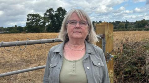 There is a woman with grey shoulder-length hair. She is wearing a grey jacket and a light green T-shirt. She has a silver necklace and hoop ear-rings. She is stood in front of a metal gate which has barbed wire on the top. Behind her is a brown field.