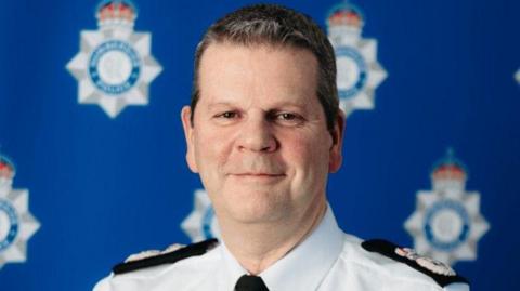 A head and shoulders shot of a man in a white shirt with police logos on his shoulders and a black tie. He is smiling at the camera. He has short brown hair and is clean shaven. There is a faded background with blurry blue police logos on it.