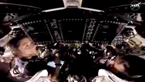 A blurry image of the crew in the spacecraft with their helmets off and sitting at the controls