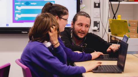 A teacher explains coding with the micro:bit to two pupils sitting at a desk