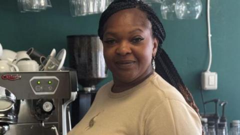 Bukola George, with long dark braided hair, tied back, standing in a cafe by a coffee machine. She is wearing a grey sweater and is looking at the camera and smiling.