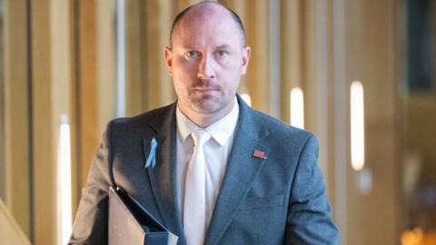 Neil Gray, who has a shaved head, looks straight ahead at the camera while walking in a wood-panelled corridor. He is wearing a grey suit with a white shirt and tie. 