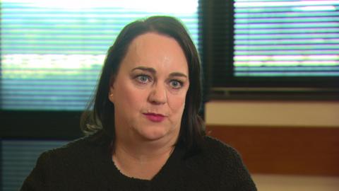 a woman looking at an off-screen interviewer. Her gaze is direct and she is wearing a black top and plum-coloured lipstick