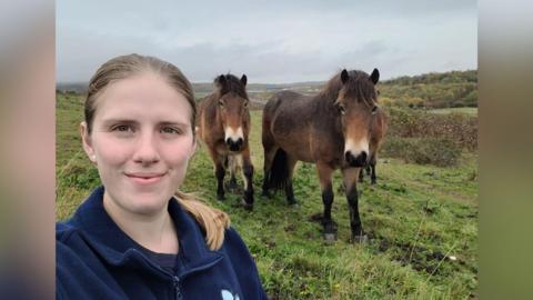 A blonde woman wearing a navy blue jumper is standing in front of three brown ponies on a field.