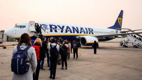 Passengers boarding a Ryanair plane. Some are standing on the tarmac while others are climbing the steps. Most are wearing backpacks.