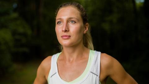 A woman with tied-back long blonde hair, wearing a white running vest. She is standing outside against a background of trees. 