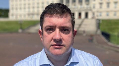 Dr David Farren, a man with dark hair, wearing a blue shirt, standing in front of Stormont Parlaiment Buildings.