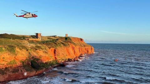 A wide shot of a helicopter flying over a cliff face and over water. The sky is blue and cloudless.