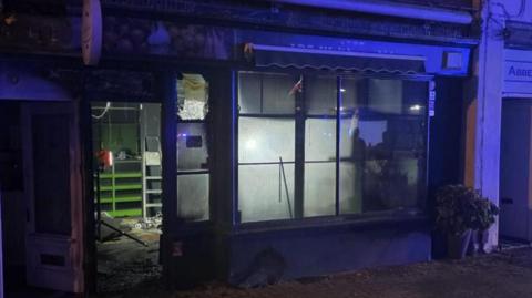 The front of a small shop. It is illuminated by the blue lights of fire engines. The windows are covered in soot from the fire and the inside is charred.