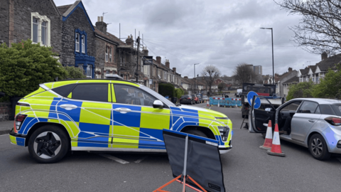 The scene of the incident in Speedwell Road, Bristol. A police car blocks the road, with another car on the right parked with the passenger door wide open. A cordon is in place on the road.