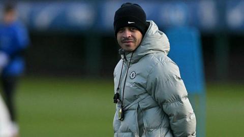Liam Rosenior, wearing a silver puffer jacket and a black woolly hat and with a whistle and stopwatch around his neck, watches a Chelsea training session 