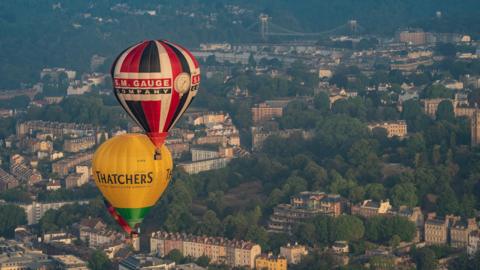 Two hot air balloons flying over Bristol. The city can be seen below, with the landscape featuring colourful properties and the Clifton Suspension Bridge as well as thick lines of trees.