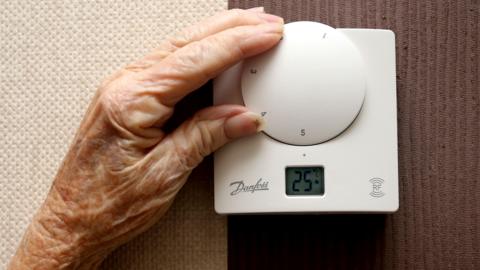 An older persons hand moves the dial on a white thermostat, the dial reads 25 degrees celcius