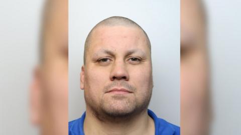 A police mug shot of a man with a shaved head and facial hair is wearing a blue collared top. The sides of the photo are blurred.