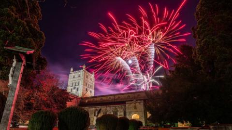 Pink, white and purple fireworks light up the dark sky behind Tewkesbury Abbey which is also lit up. The image is taken from ground level.