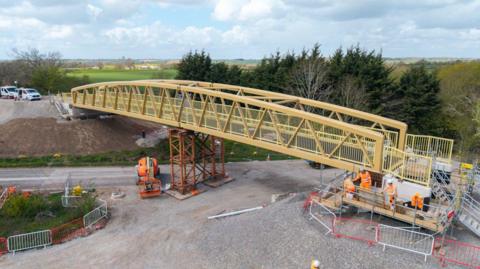 A pedestrian bridge is being installed over a road, with construction workers and equipment below.