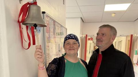 A woman with a navy scarf on her head and a green shirt. She is ringing a silver bell and standing next to a man wearing a red shirt and black jacket.