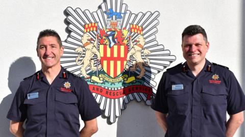 Chief Fire Officer Keith Carruthers and deputy chief Stephen Kennedy are standing with their arms behind their back and smiling brightly at the camera. There white and red fire service crest is mounted on the white wall behind them. They are both wearing dark blue fire service short sleeved button-up shirts with light blue name badges.