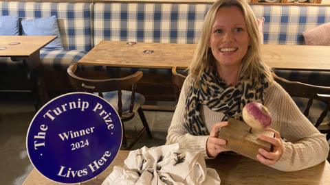 A woman smiles at the camera holding a turnip with a nail through it. She is sitting next to a sign saying 'Turnip Prize winner' and a white crumpled up shirt with tacks in it.