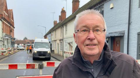 Dave Tucker, wearing glasses and a brown coat stood in front of a red and white barrier on a closed road