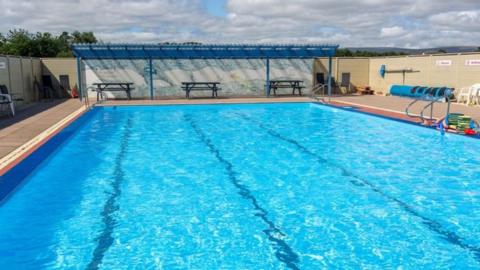 Lengthways view of a swimming pool full of water, but with no swimmers. There are benches on a partially-covered paved area at one end, and to the left a rolled up blue tarpaulin cover and some swimming aids.