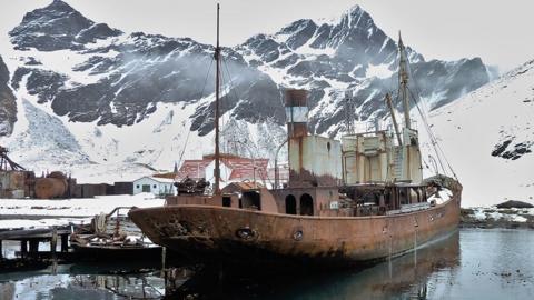 A rusting steam trawler in a rundown harbour with snow-covered mountains in the background. The hull and superstructure is covered in rust. In the background are huts and rusting machinery surrounded by snow.
