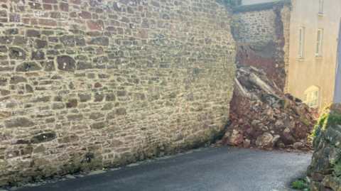 A high brown wall with a section next to a cream house which has collapsed into the road. The red mortar can be seen in the debris