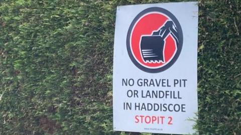 A sign mounted on a leylandii hedge. It depicts a logo - a black circle, infilled with red and a black digger bucket. The words state: "No gravel pit or landfill in Haddiscoe. Stopit 2"