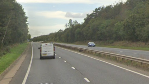 View of a dual carriageway standing on the westbound route. A van is travelling westbound on the left side of the two lanes. A car is travelling towards on the right going eastbound. Trees are lined either side of the A47.