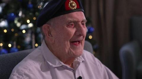 An elderly veteran wearing a navy beret with a green and gold logo at the front. He is smiling with his mouth open and wearing a long-sleeved light pink shirt. He is sat in a grey chair and the background is blurred but you can see fairy lights from a Christmas tree.