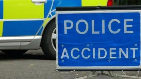 A blue sign on the road which says POLICE ACCIDENT in white writing. It is on the road next to a police car. 