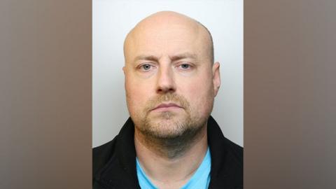 A police issue picture of Richard Mills, he had a blonde beard wears a black jacket and a blue t-shirt. 