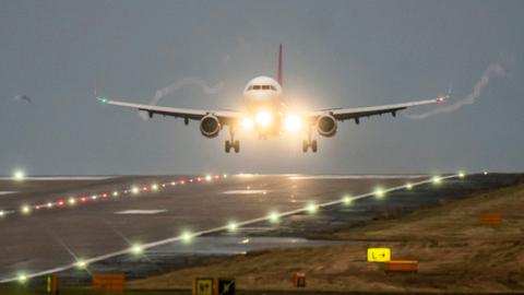 A plane comes in to land at Leeds Bradford Airport in dusky conditions. Lights can be seen at the front of the plane and along the runway.
