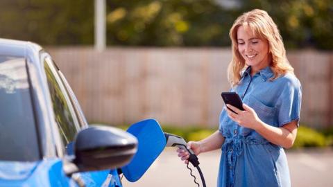 A woman charges an electric car while using an app on her phone to monitor the battery level. She is wearing a blue summer dress and there is a fence and trees behind her. The blue car is parked in a road.