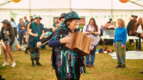 A woman wearing colourful clothes is stading in the middle of a large marquee playing a wooden accordian. Her hat is green and her jacket has pieces of fabric like streamers attached in the same green, purple, white and black. There are other similarly dressed people behind her and not infocus in the background. 