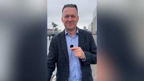Scottish Conservative leader Russell Findlay smiles at the camera as he stands in front of the Clyde river with buildings in the background. He has grey hair, blue eyes and is wearing a blue shirt and navy suit jacket.