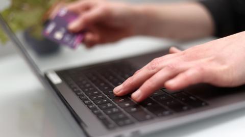 A hand is seen typing on a grey laptop. The other hand is holding a purple bank card.